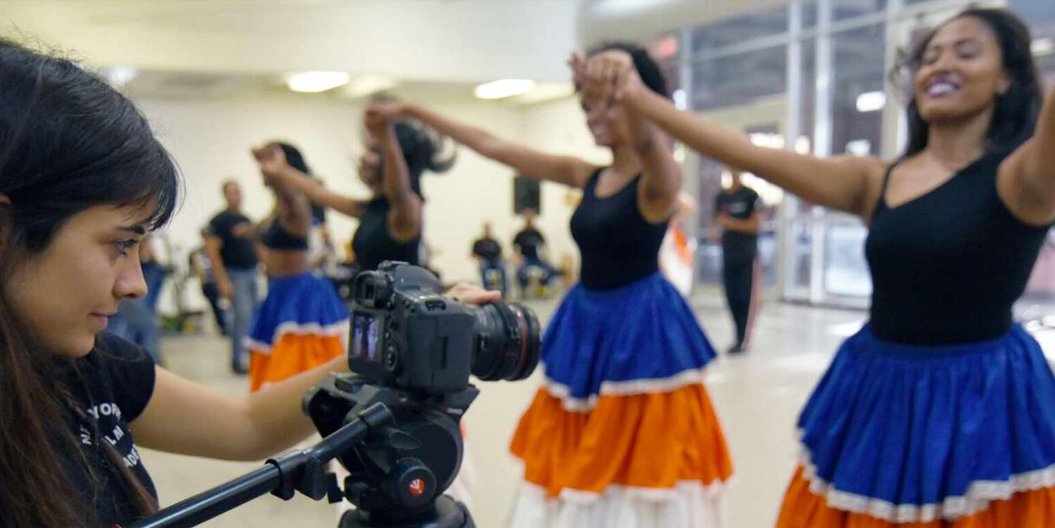Woman videotaping women dancing in blue and orange skirts