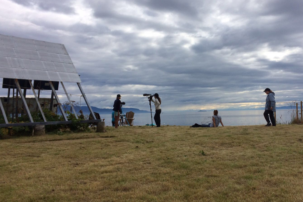 People videotaping ocean with grass in front and mountains in background
