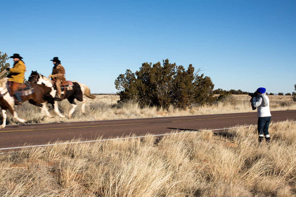 Woman photographing men riding on horses down desert road