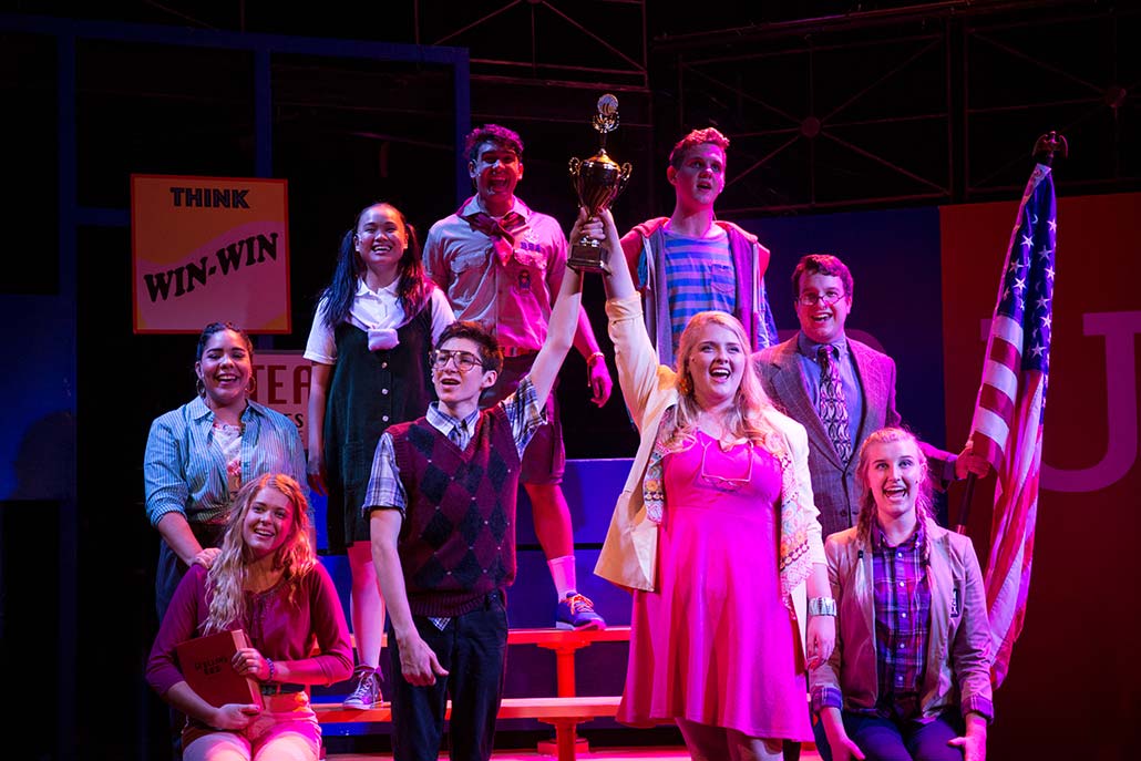 Spelling Bee student ensemble sings with big trophy on belachers.