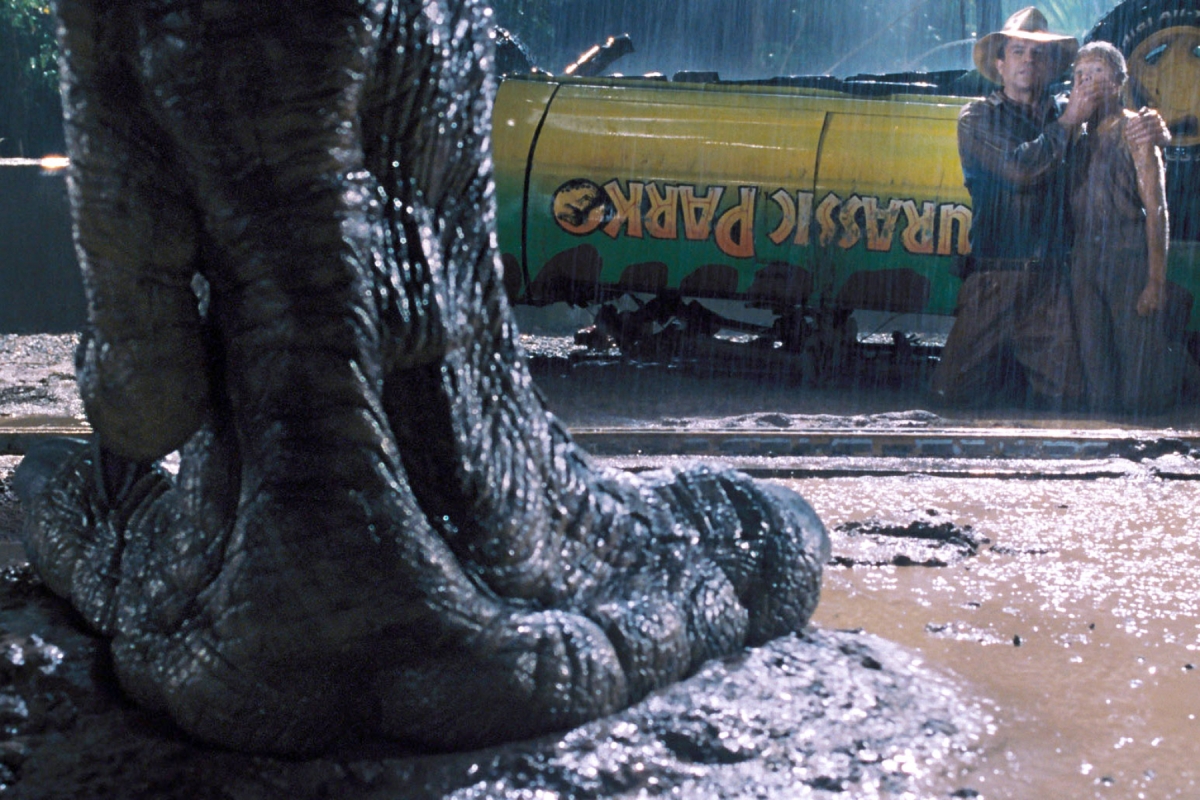 Scene from the film Jurassic Park. Large dinosaur foot stands in front of overturned car and man holds his hand over a child's mouth to keep them quiet